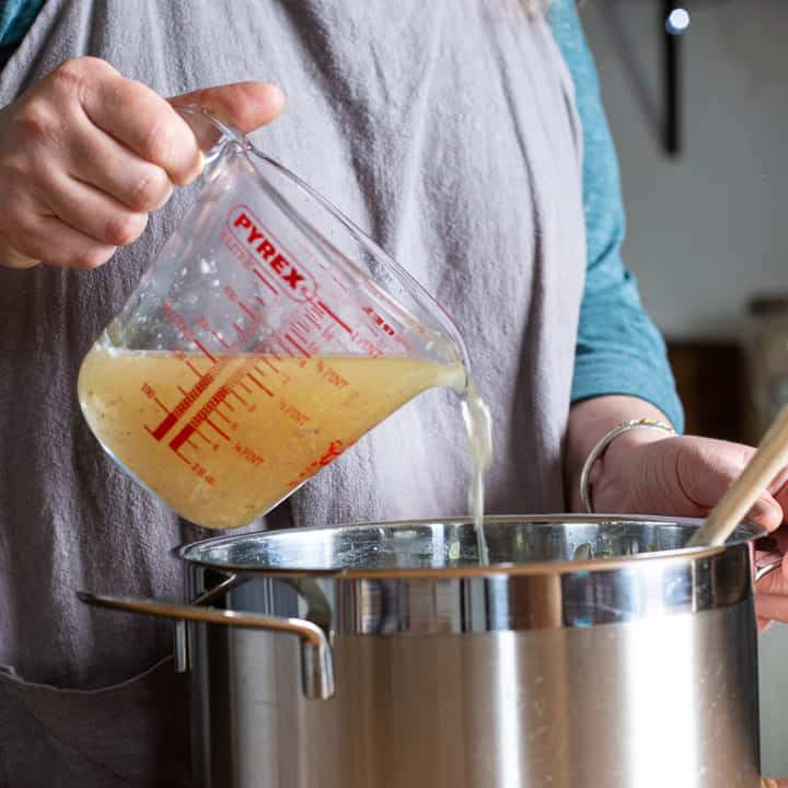 woman in grey pouring stock from a glass jug into a large silver saucepan