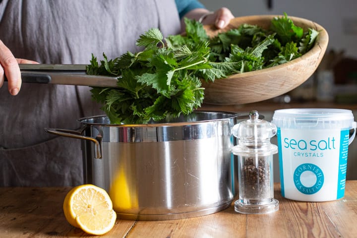 woman in grey using silver tongs to move stinging nettles from a wooden bowl into a large silver saucepan