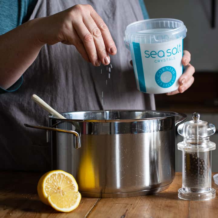 woman in grey sprinkling sea salt from a large blue and white tub into a silver saucepan of soup on a wooden kitchen counter