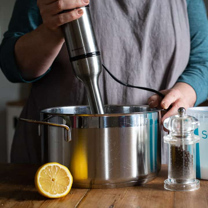 womans hands holding an immersion blender over a large silver pan with half a lemon and a pepper mill in front