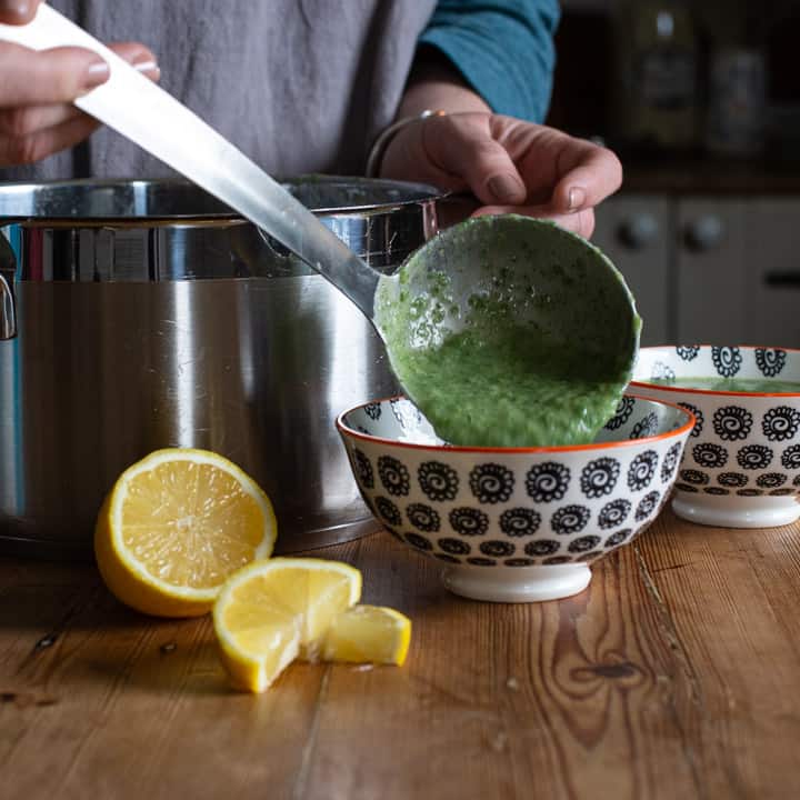 womans hands spooning bright green soup into smal white bowls with a silver ladle on a wooden kitchen counter