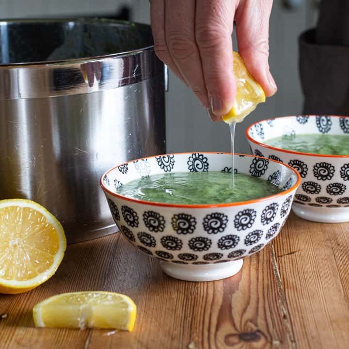 womans hand squeezing a wedge of lemon into a small white and black bowl full of green nettle soup