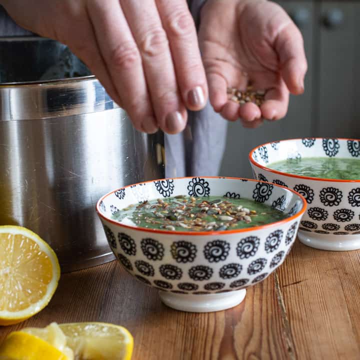 woman sprinkling mixed seeds onto a bowl of bright green soup next to lemon slices and a silver saucepan