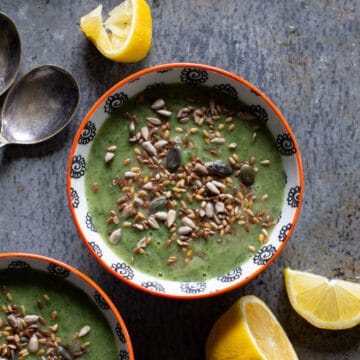 2 orange and white bowls filled with a green soup sprinkled with mixed seeds on a grey metal background with soup spoons and lemon slices