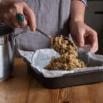 Woman scooping flapjack mix into a lined metal baking dish