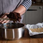 Woman scooping softened dates from a small silver pan into a baking tin with a wooden spoon