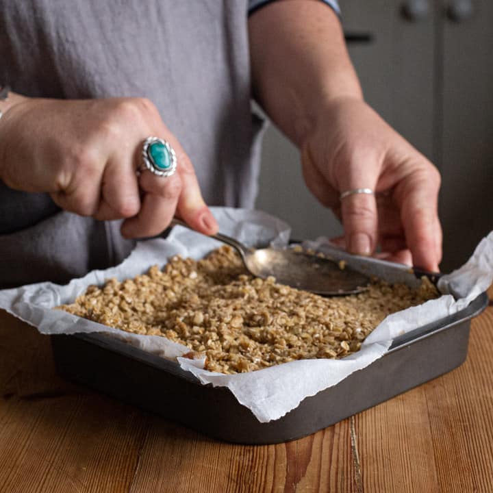 Woman pressing oat bar mixtures into a grey metal baking tin lined with white baking paper