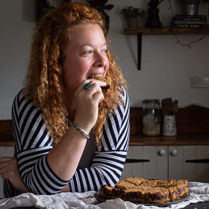 woman with red hair in stripey top leaning on wooden kitchen counter eating fresh date flapjack