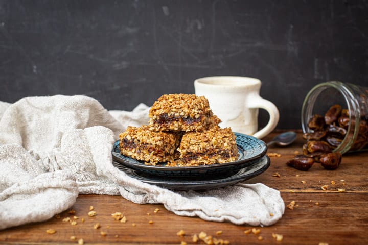 Plates of best flapjacks on a cloth sat on a table, mug behind them and dates spilling from a jar
