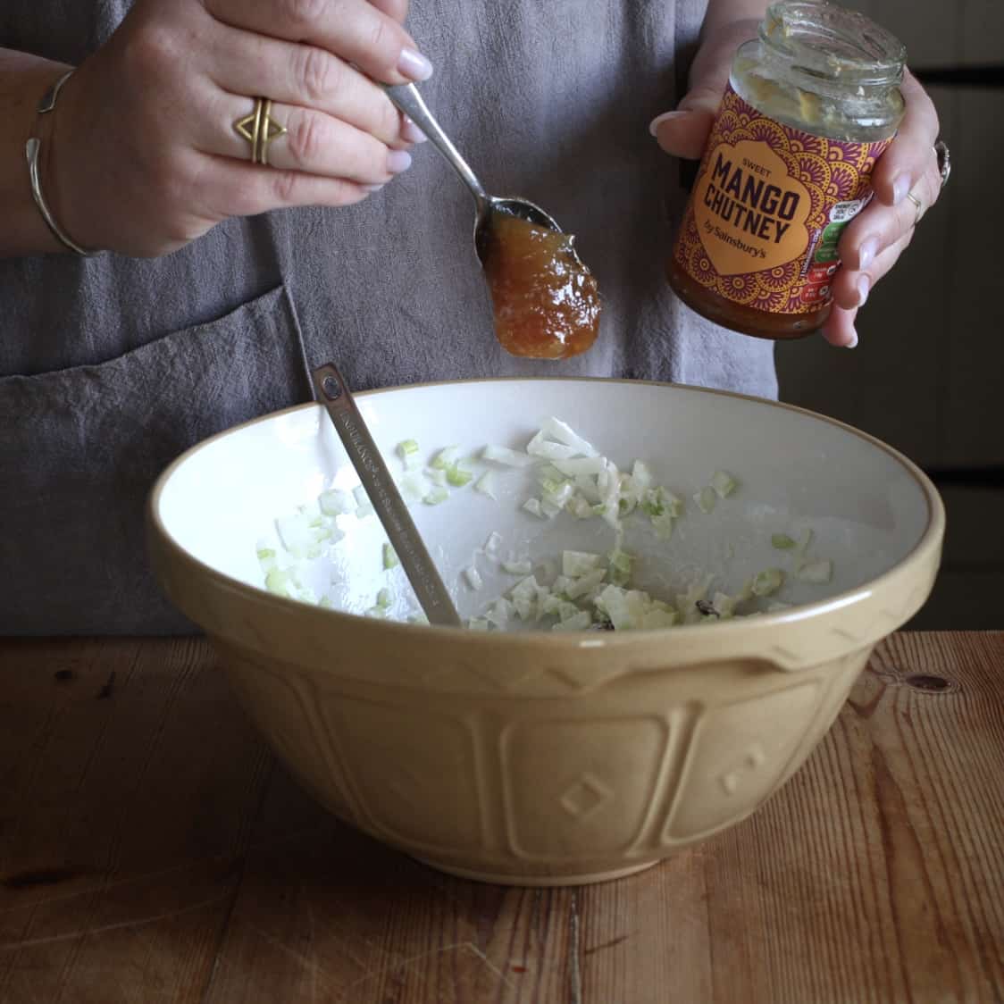 Woman dropping a spoonful of mango chutney into a brown mixing bowl from a silver teaspoon