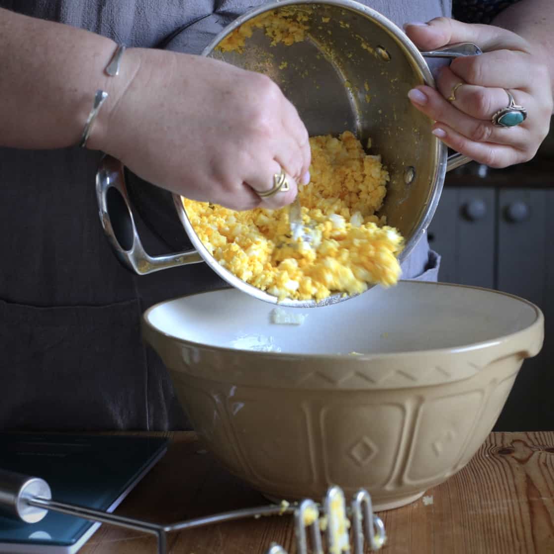 Womans scraping mashed hard boiled eggs from a silver saucepan into a brown mixing bowl
