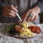 Woman sliding coronation egg salad from a silver dessert spoon onto a plate of brightly coloured salads