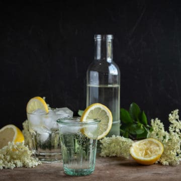 glass bottle of homemade elderflower cordial with lemons, flowers and glasses piled high with cordial and ice