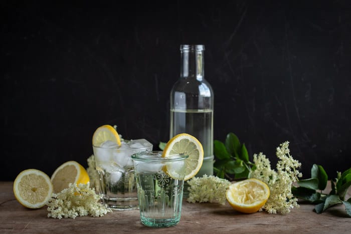 glass bottle of homemade elderflower cordial with lemons, flowers and glasses piled high with cordial and ice