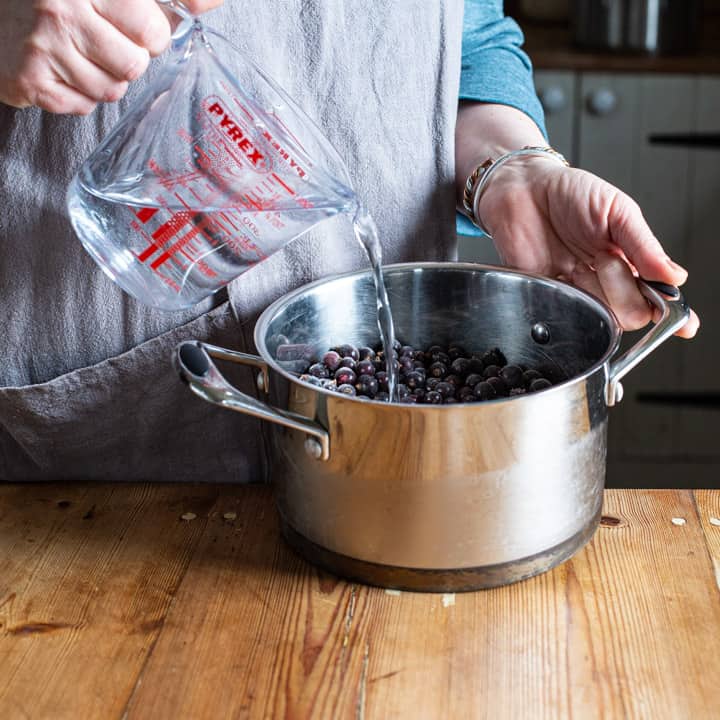 womans hands pouring water from a glass jug into a silver pan of blackcurrants