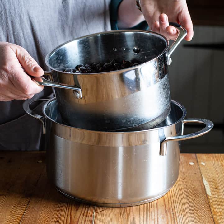 womans hands holding a small silver saucepan over a large silver saucepan for a makeshift bain maire