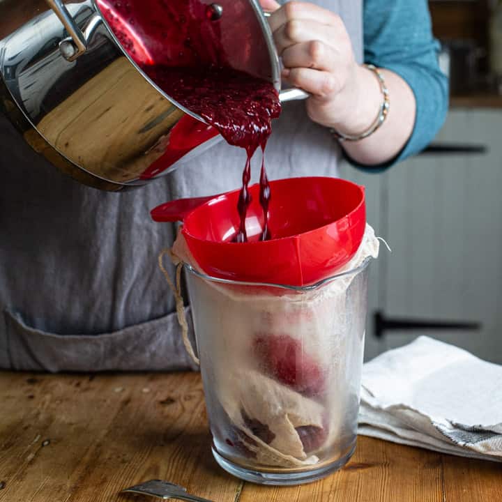 womans hands tipping lightly stewed balckcurrants from a silver saucepan into a muslin bag over a glass jug