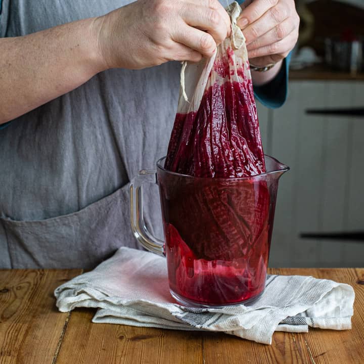 womans hands draining the juice from a bag of blackcurrants into a glass jug on a wooden kitchen counter