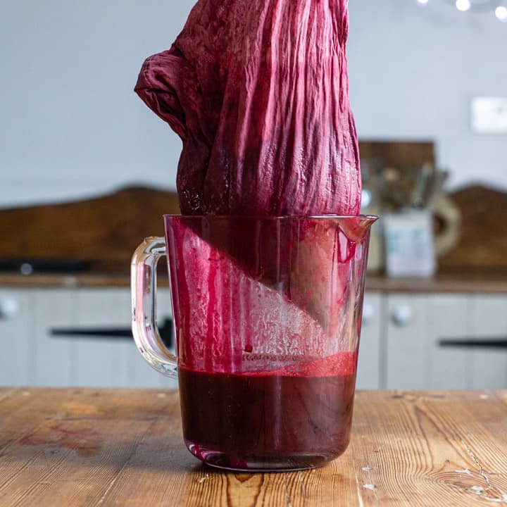 wooden counter with a glass jug half filled with blackcurrant juice and muslin bag of blackcurrants draining into it
