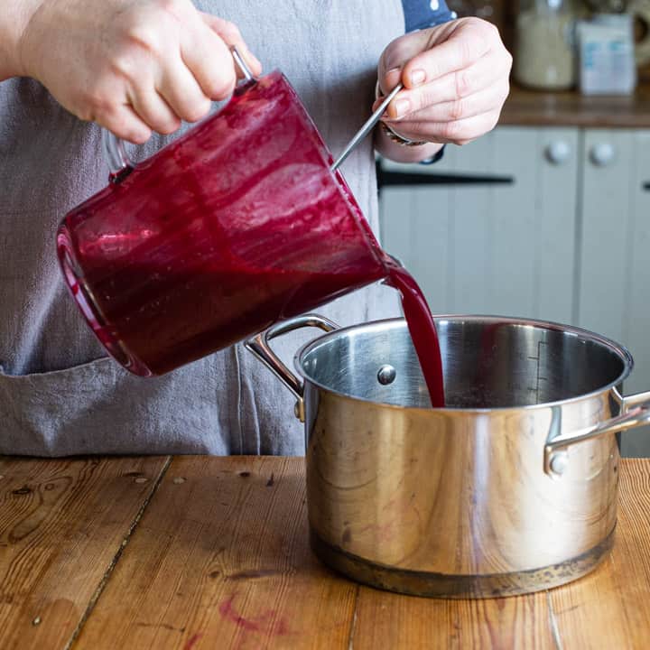 womans hands pouring blackcurrant juice from a glass jug into a silver saucepan on a wooden counter