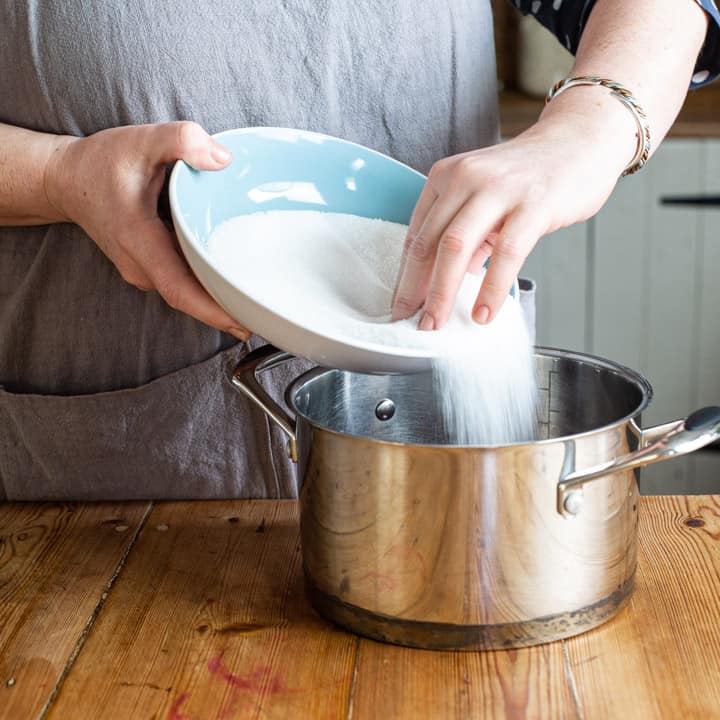 womans hands tipping white sugar from a blue bowl into a silver saucepan of blackcurrant juice