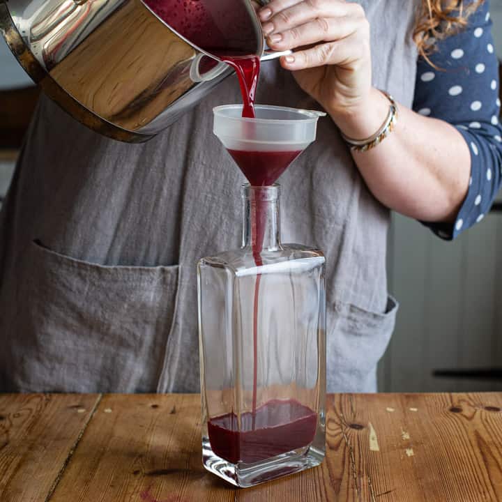 woman in grey apron pouring blackcurrants syrup from a silver pan, into a plastic funnel over a glass bottle