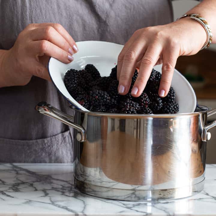 womans ahnds tipping fresh blackberries from a white bowl into a silver saucepan