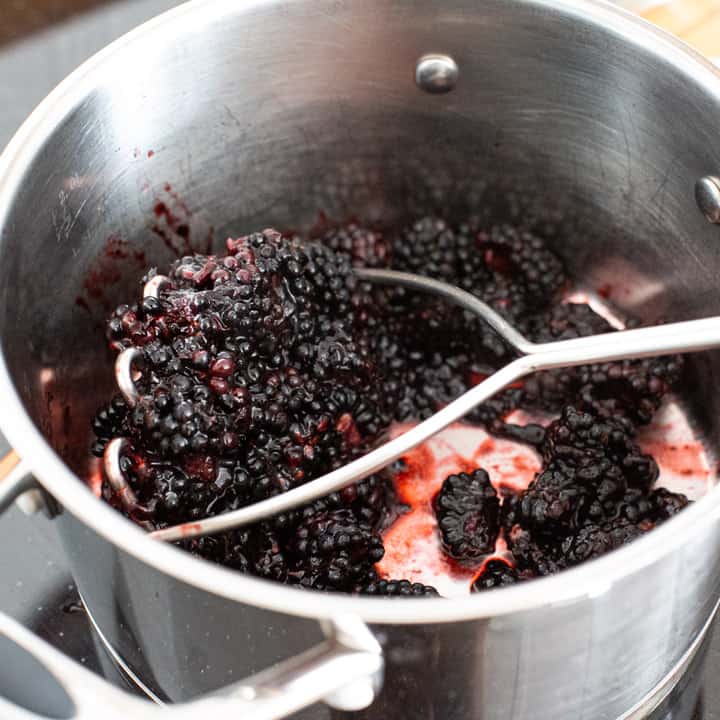 inside a silver saucepan filled fresh blackberries getting squashed with a silver potato masher