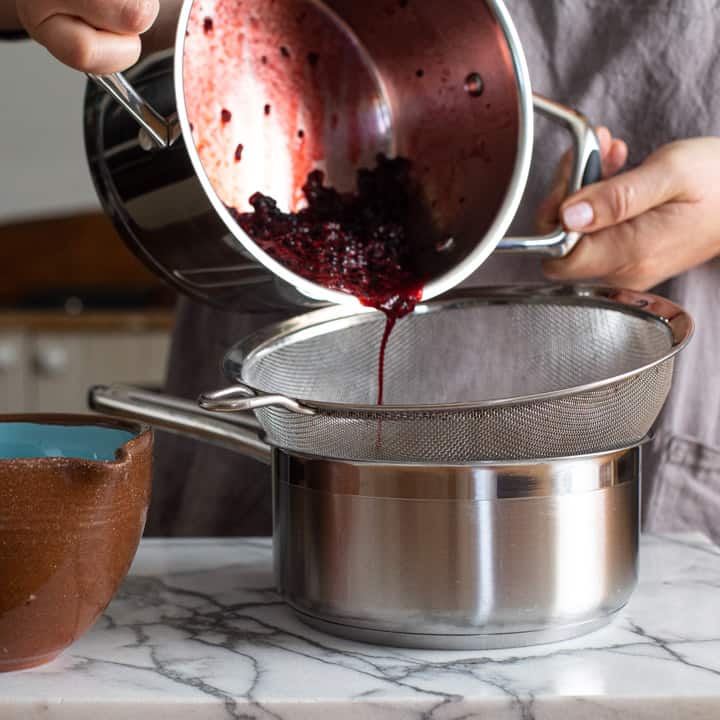 womans hands pouring cooed blackberries into a silver sieve over a silver saucepan