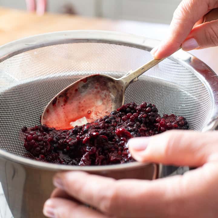 womans hands squashing cooked blackberries in a silver sieve to extract the juice