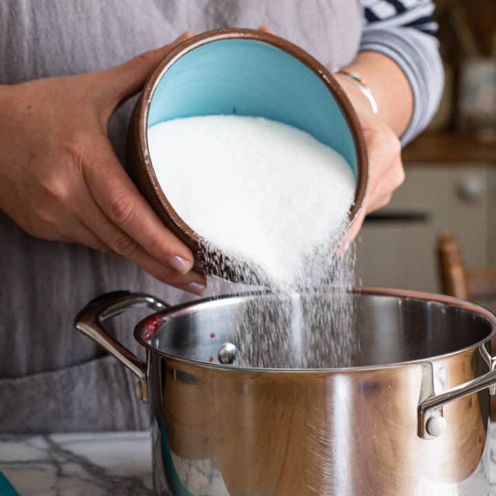 womans hands tipping a blue bowl of sugar into a silver pan of blackberry juice