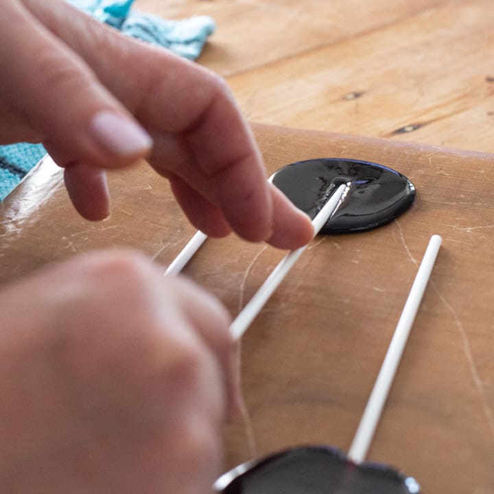 womans hands pushing a white paper lollipop stick into a circle of hot blackberry candy on a brown baking sheet 