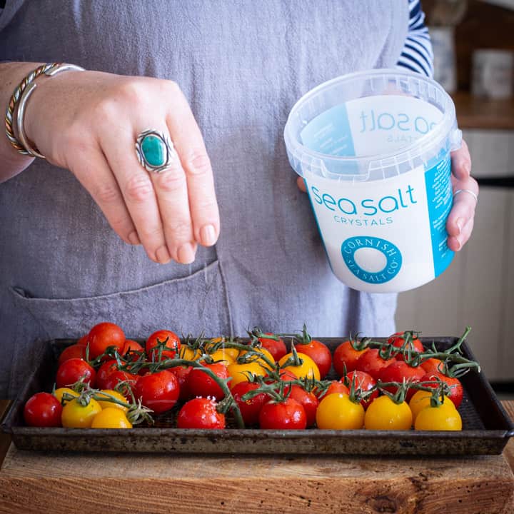 womans hands sprinkling sea salt on a tray of red and yellow cherry tomatoes for roasting