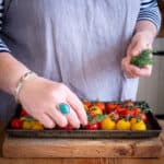 womans hands placing fresh herbs on a tray of red and yellow cherry tomatoes for roasting