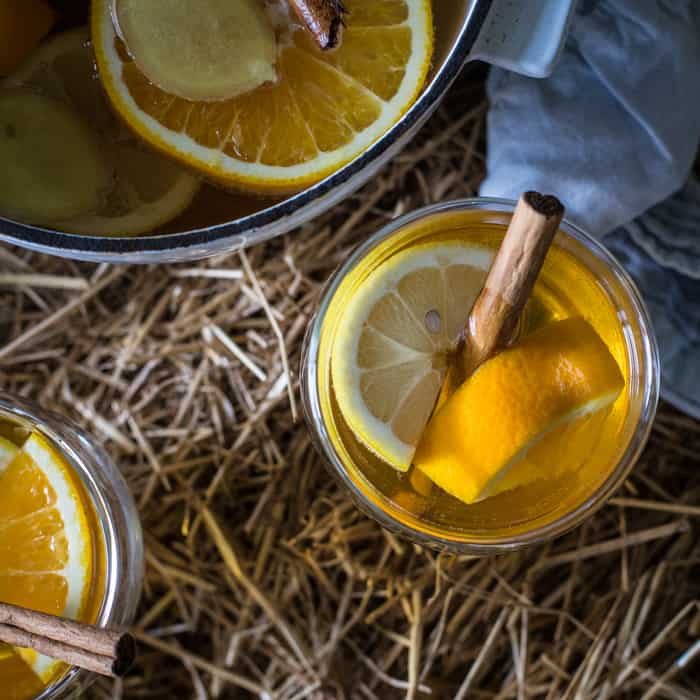 Two full glasses and a pan of mulled cider on straw bale with slices of lemon and cinnamon sticks