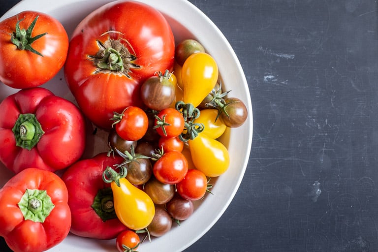 black background with white bowl full of fresh yellow, orange and red tomatoes