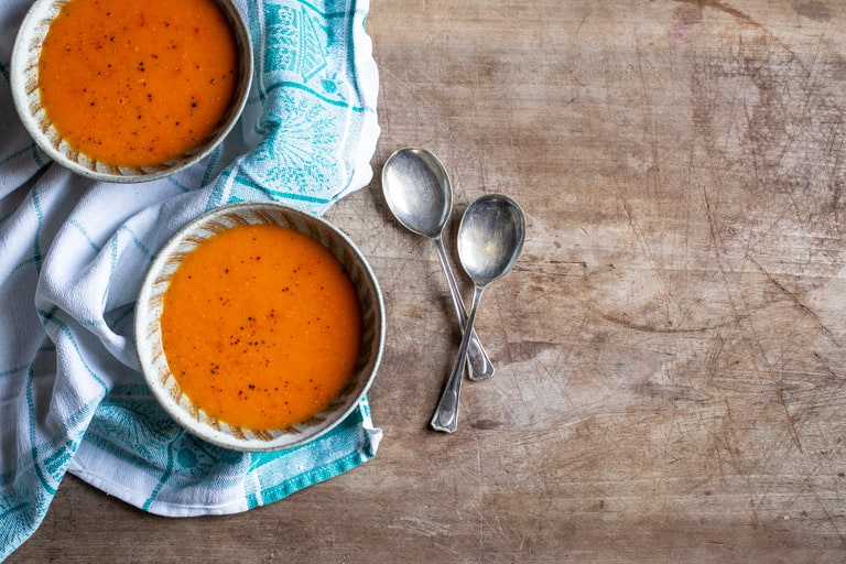 wooden backgrouns with 2 bowls of red pepper tomato soup on a blue and white cloth with 2 spoons