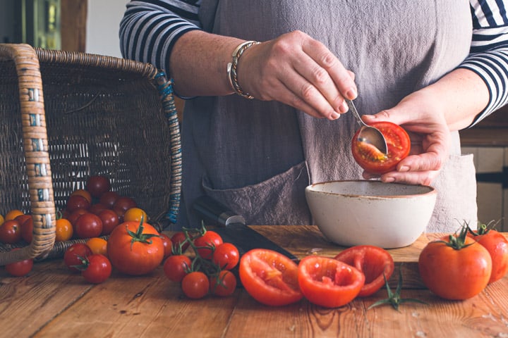 womans hands scooping seeds out of red tomato on wooden chopping board