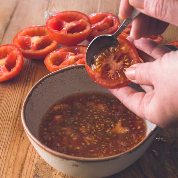 womans hands scooping seeds out of red tomato on wooden chopping board