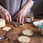 womans hands on wooden kitchen bench spreading fresh hot oatcakes with chutney surrounded by baking mess and cheddar cheese