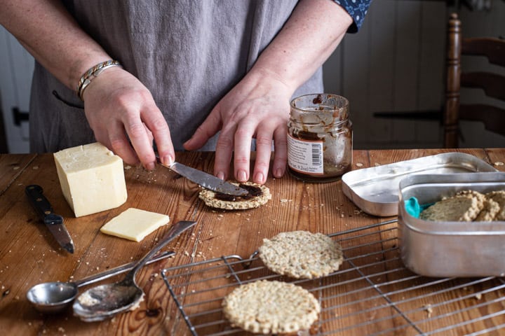 womans hands on wooden kitchen bench spreading fresh hot oatcakes with chutney surrounded by baking mess and cheddar cheese