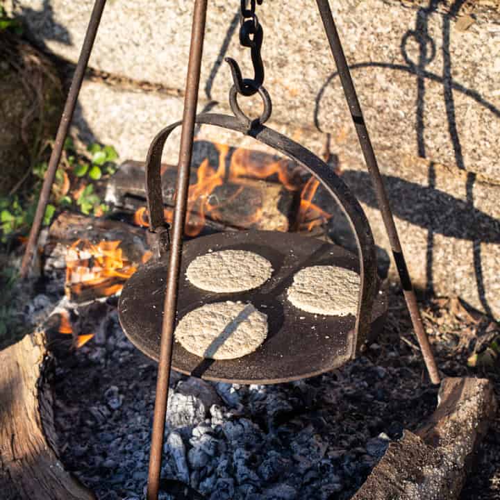 cast iron griddle hanging from a hook over a campfire with 3 homemade oatcakes cooking on the hot plate 