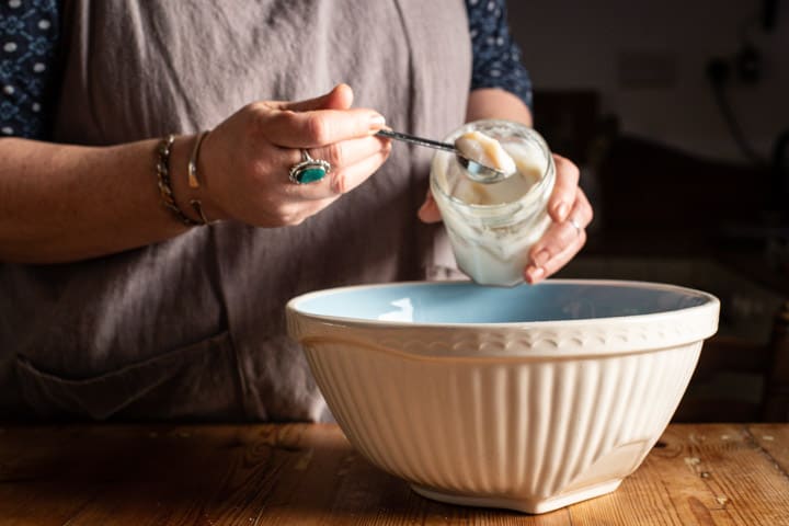 womans hands spooning lard from a glass jar into a white mixing bowl of oatcake ingredients