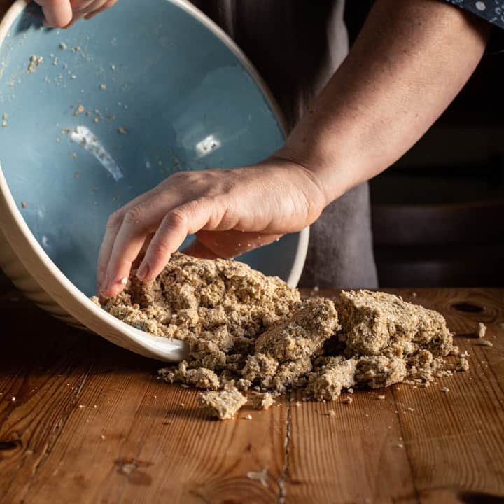 womans hands tipping crumbly oatcake dough from a blue and white bowl onto a wooden kitchen counter