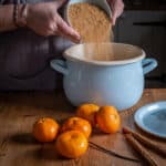 rustic kitchen counter with womans hands pouring a bowl of brown sugar into a pale blue enamel saucepan with satsumas and cinnamon sticks on the counter