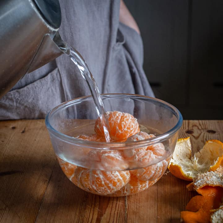 wooden kitchen counter with glass bowl of peeled satsumas and boiling water being poured from a kettle into the bowl