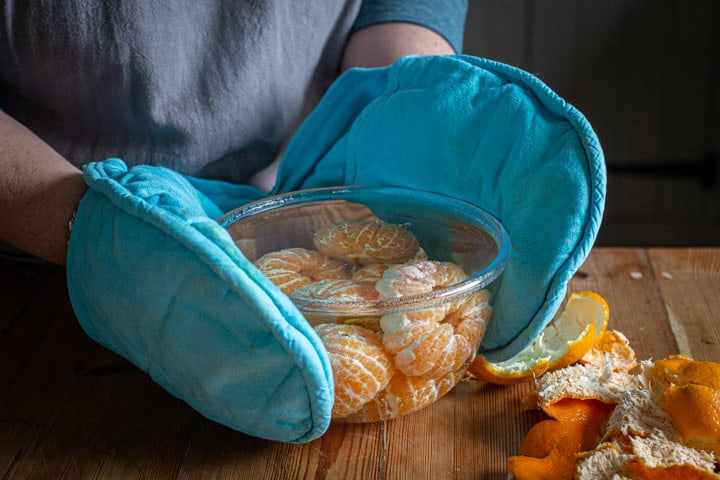 blanched whole satsumas in a glass bowl being held by hands in blue oven gloves