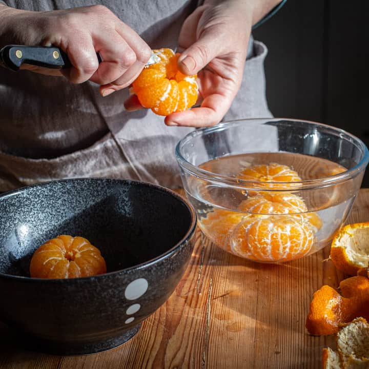womans hands gently scraping the pith from a whole poached satsuma over a rustic wooden kitchen counter