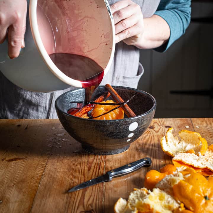 womans hands pouring red wine syrup from a large blue saucepan over a black bowl containing poached whole satsumas on a rustic wooden kitchen counter