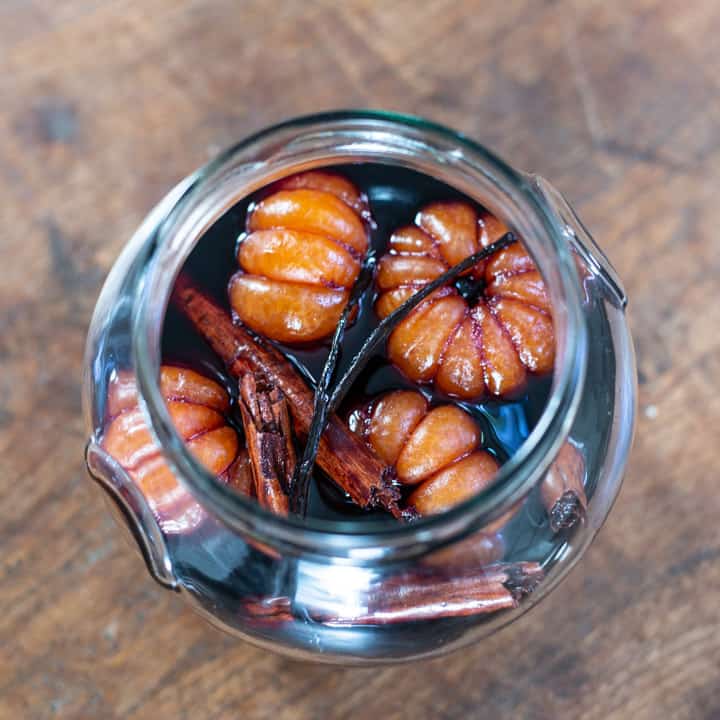 large glass jar on wooden background background with whole poached satsumas, red wine syrup, cinnamon sticks and a vanilla pod